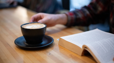 Person holding a cup of coffee next to an open book on a wooden table
